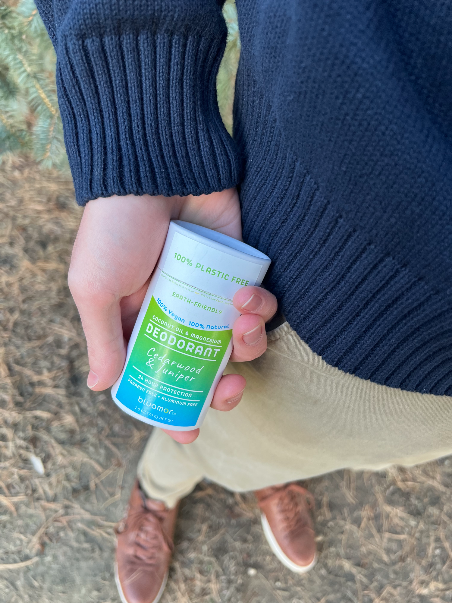 A person in a dark sweater holds a plastic-free deodorant stick labeled "Cedarwood & Juniper" outdoors.