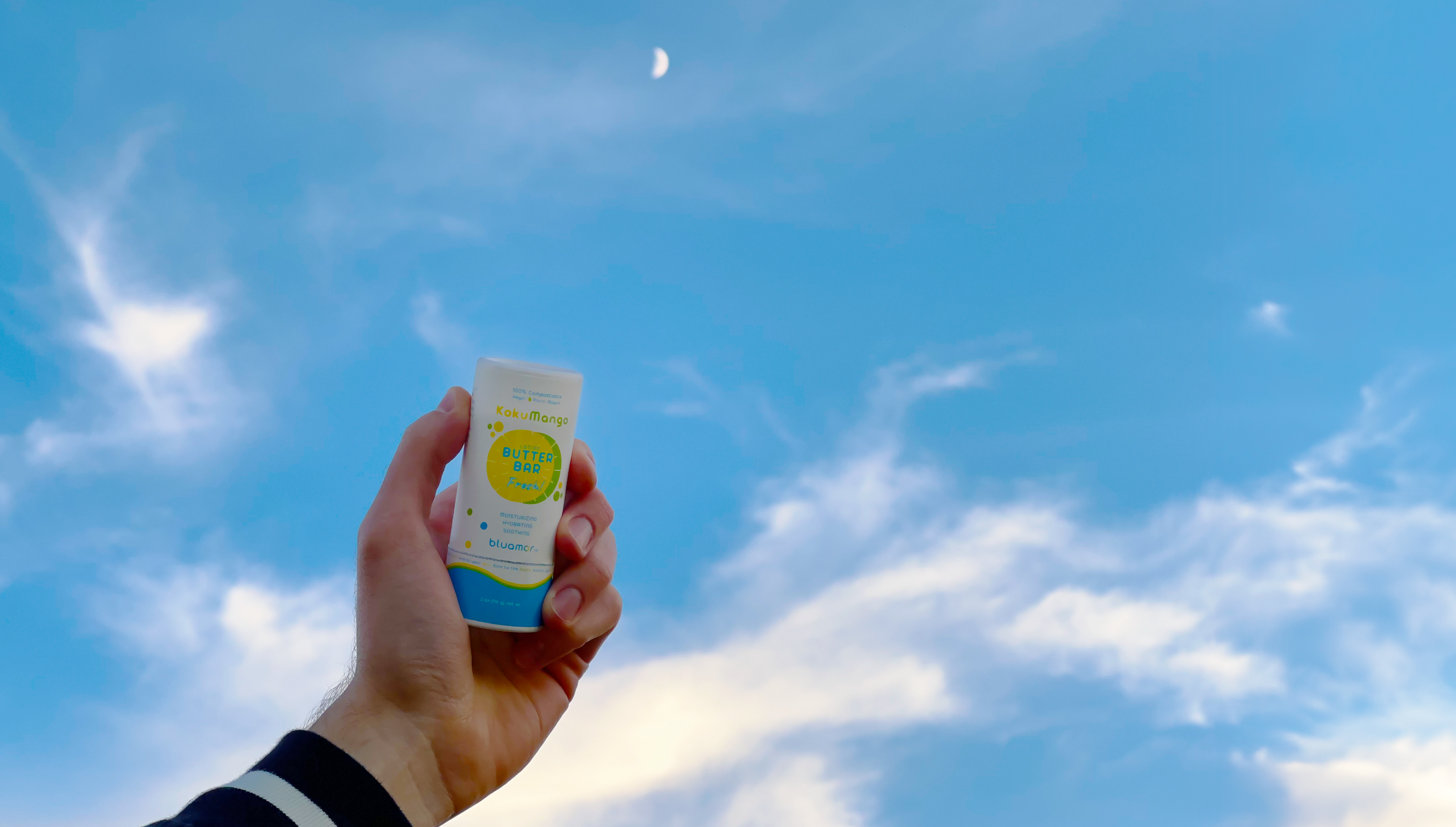 A hand holds a bluamor KokuMango Butter Bar Fresh against a bright blue sky with wispy clouds and a crescent moon in the background.