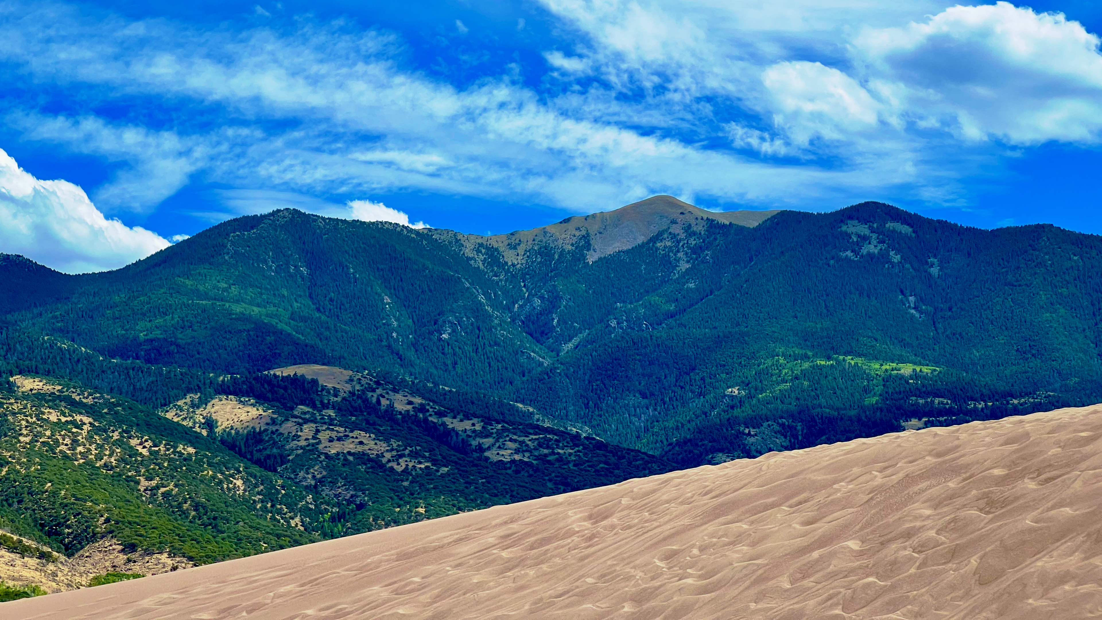 A sandy dune in the foreground contrasts with lush green mountains and a bright blue sky, dotted with fluffy white clouds.