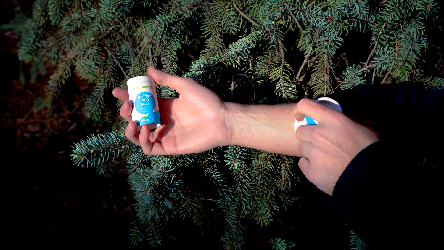 A person applies bluamor lotion butter bar, Woods, to their arm while holding the container, surrounded by green pine branches.