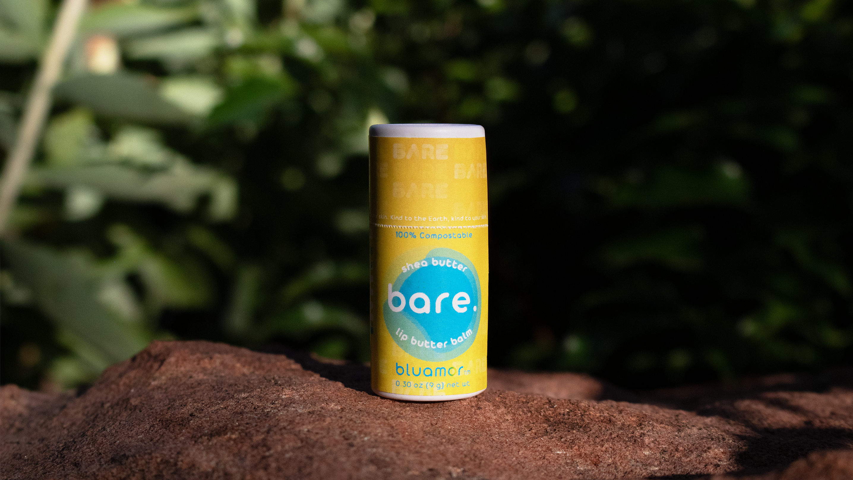 A yellow tube of bluamor lip butter balm bare. Shea butter lip balm sits on a stone, surrounded by green foliage, emphasizing its eco-friendly branding.