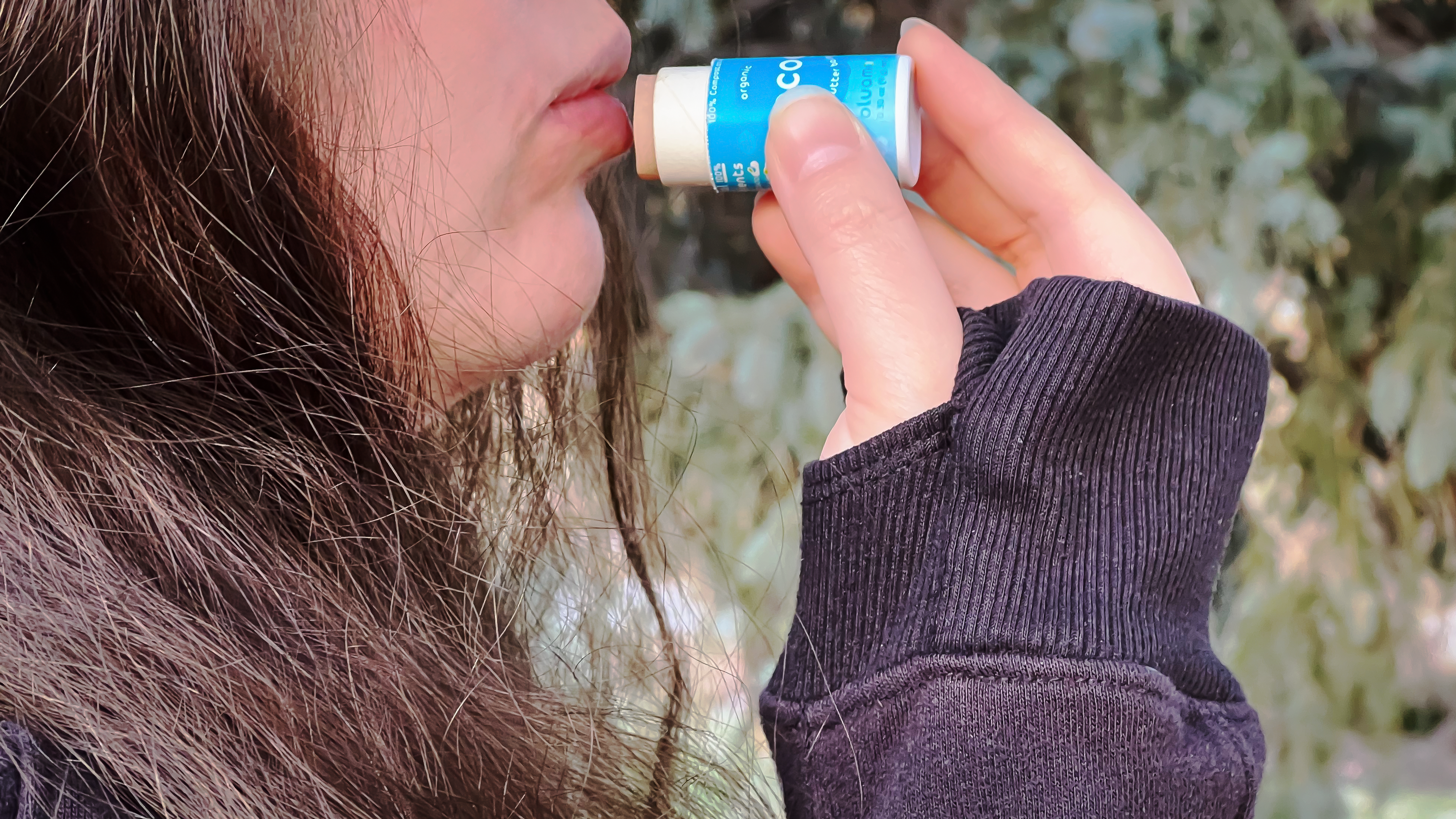 A close-up of a woman applying lip balm to her lips, wearing a black sweater and surrounded by blurred greenery.