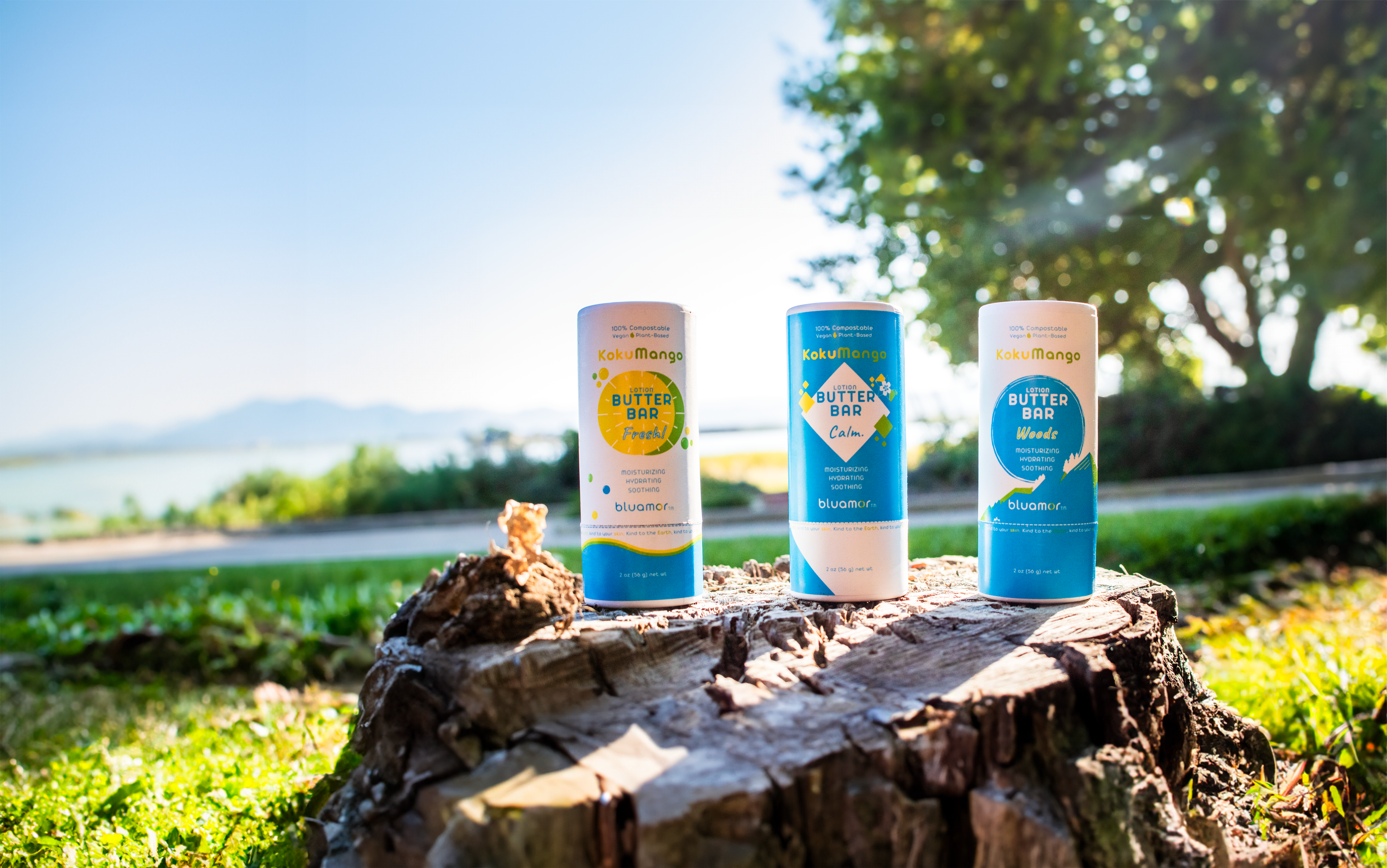 Three colorful bluamor KokuMango Lotion Butter Bars labeled "Fresh," "Calm," and "Woods," placed on a wooden stump with a scenic background.